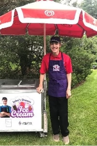 John is standing next to his ice cream stand with a red umbrella